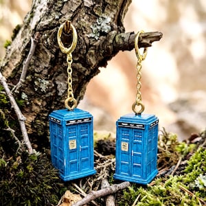 May include: A pair of drop earrings featuring miniature blue police box charms suspended from gold-toned metal chains and hoop findings. Each blue box charm displays a small white sign with black text on the front door panel and a black roof.