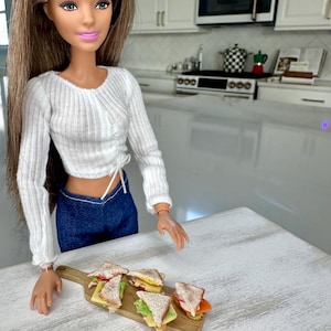 May include: A doll wearing a white long-sleeved top and blue jeans stands near a wooden cutting board with miniature sandwiches. The sandwiches have various fillings, including cheese, lettuce, and tomato. The doll has long, light brown hair and pink lipstick.