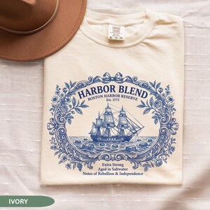 May include: An ivory t-shirt featuring a blue graphic of a sailing ship surrounded by an ornate floral frame. The text reads HARBOR BLEND BOSTON HARBOR RESERVE Est. 1773, Extra Strong, Aged in Saltwater, Notes of Rebellion & Independence.