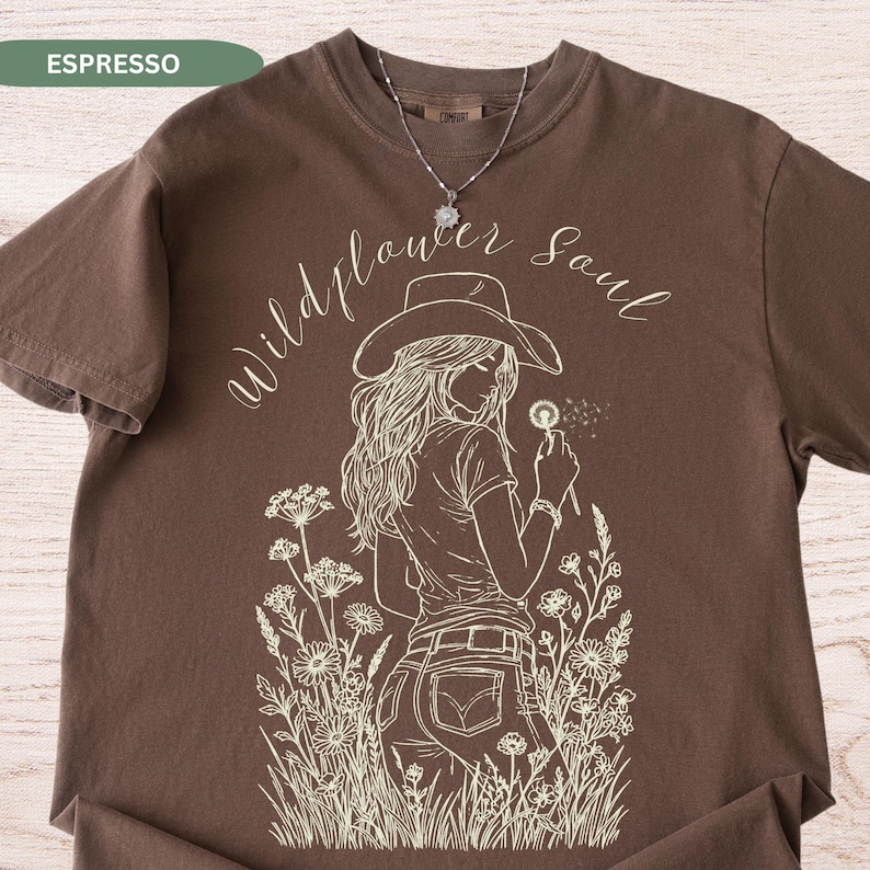 Puede incluir: Camiseta marr&oacute;n con un dibujo lineal blanco de una persona con sombrero de vaquero sosteniendo un diente de le&oacute;n. Las palabras "Wildflower Soul" est&aacute;n escritas en cursiva encima de la imagen. Se ve un collar plateado.