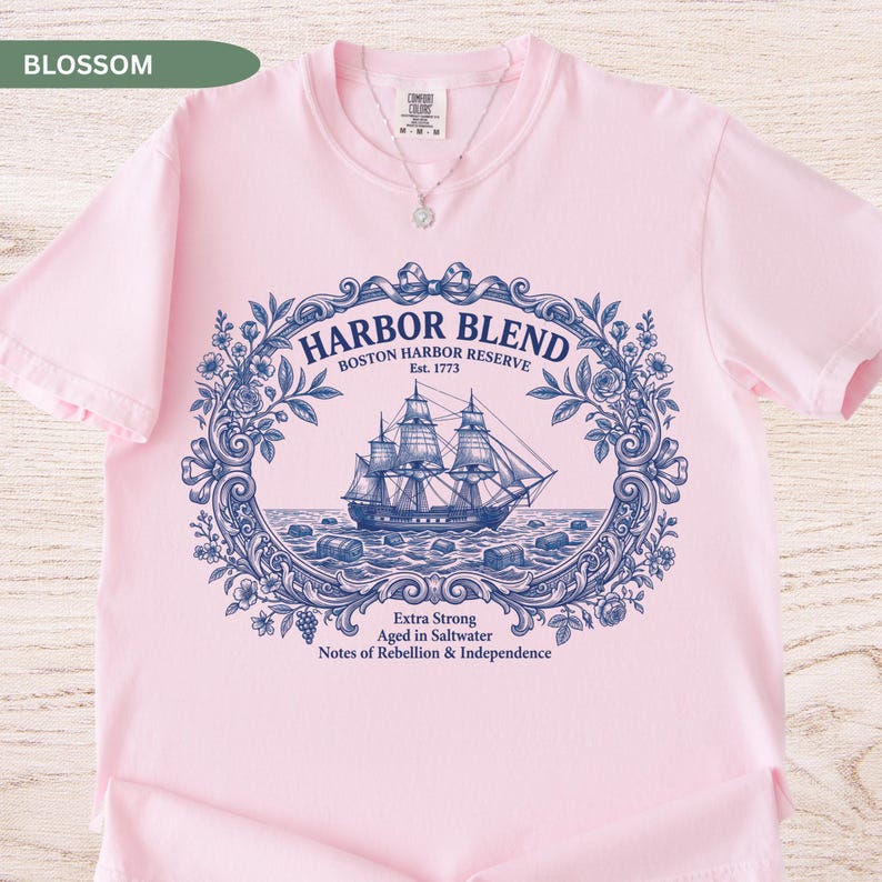 May include: A light pink t-shirt featuring a dark blue graphic of a sailing ship inside an ornate floral frame. The text reads Harbor Blend, Boston Harbor Reserve, Est. 1773, Extra Strong, Aged in Saltwater, Notes of Rebellion & Independence.