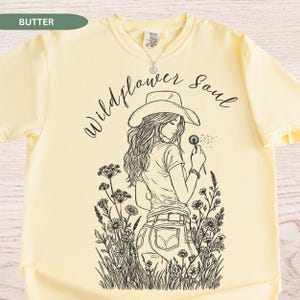 Puede incluir: Camiseta de color mantequilla con un gr&aacute;fico negro de una mujer con sombrero de vaquero soplando un diente de le&oacute;n, rodeada de flores silvestres. El texto "Wildflower Soul" est&aacute; escrito encima. Se ve un collar plateado.
