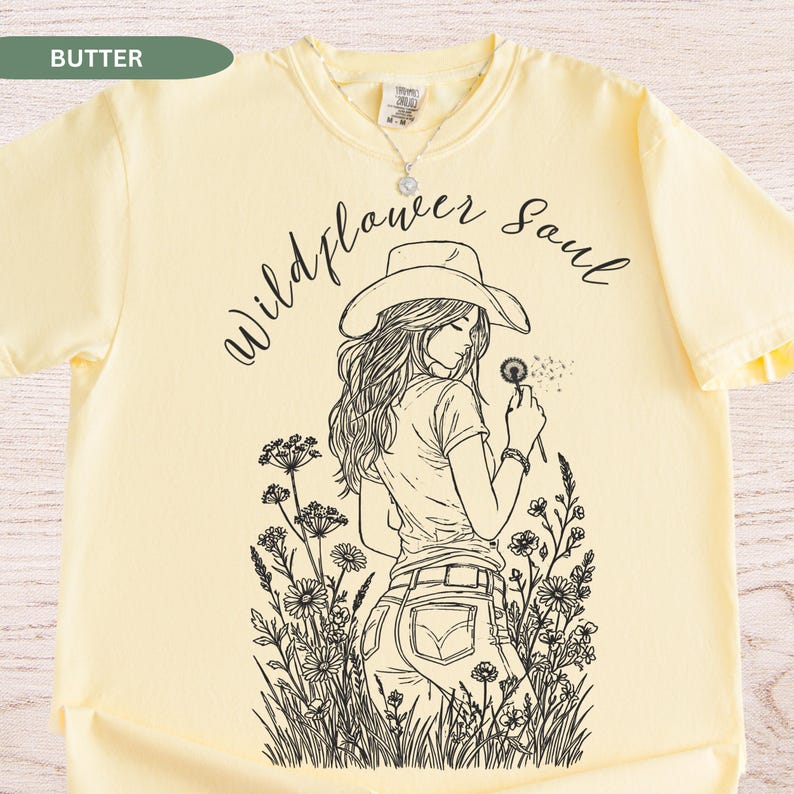 Puede incluir: Camiseta de color mantequilla con un gr&aacute;fico negro de una mujer con sombrero de vaquero soplando un diente de le&oacute;n, rodeada de flores silvestres. El texto "Wildflower Soul" est&aacute; escrito encima. Se ve un collar plateado.