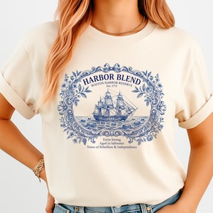 May include: A cream-colored t-shirt featuring a blue graphic print of a sailing ship centered within an ornate floral frame. The text reads HARBOR BLEND BOSTON HARBOR RESERVE Est. 1773, Extra Strong, Aged in Saltwater, Notes of Rebellion & Independence.