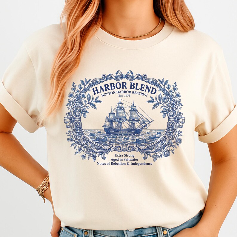 May include: A cream-colored t-shirt featuring a blue graphic print of a sailing ship centered within an ornate floral frame. The text reads HARBOR BLEND BOSTON HARBOR RESERVE Est. 1773, Extra Strong, Aged in Saltwater, Notes of Rebellion & Independence.