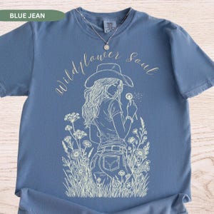 Puede incluir: Camiseta azul vaquero con un gr&aacute;fico blanco de una mujer con sombrero de vaquero sosteniendo un diente de le&oacute;n. Las palabras "Wildflower Soul" est&aacute;n escritas en cursiva sobre el gr&aacute;fico. La camiseta tiene cuello redondo y mangas cortas.