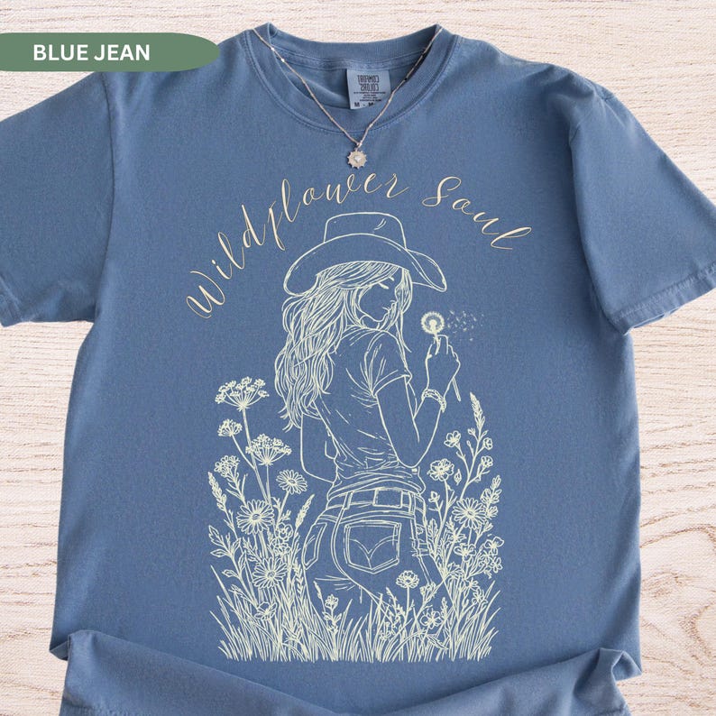 Puede incluir: Camiseta azul vaquero con un gr&aacute;fico blanco de una mujer con sombrero de vaquero sosteniendo un diente de le&oacute;n. Las palabras "Wildflower Soul" est&aacute;n escritas en cursiva sobre el gr&aacute;fico. La camiseta tiene cuello redondo y mangas cortas.