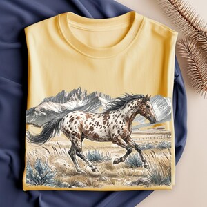 May include: A pale yellow t-shirt featuring a graphic illustration of a spotted horse running through a grassy field. In the background, there are detailed mountains under a clear sky. The artwork uses muted earth tones and shades of gray and white.