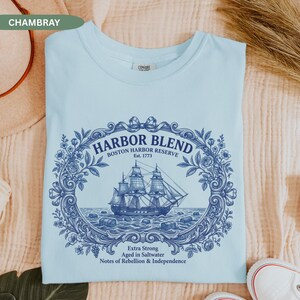 May include: A light blue t-shirt featuring a dark blue graphic of a sailing ship centered within an ornate floral frame. The text reads HARBOR BLEND BOSTON HARBOR RESERVE Est. 1773, Extra Strong, Aged in Saltwater, Notes of Rebellion & Independence.