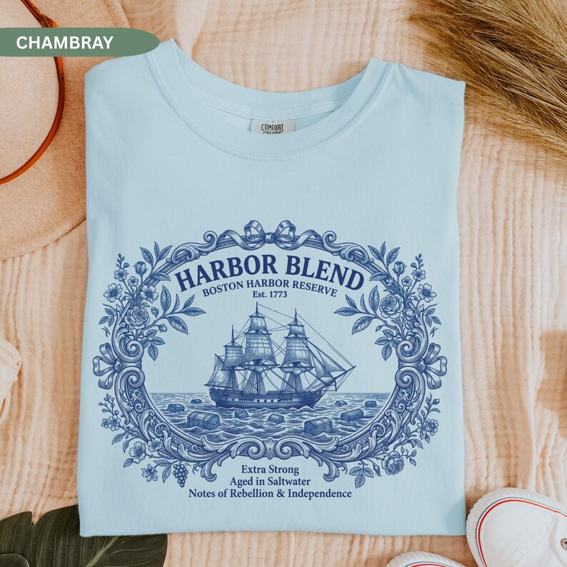 May include: A light blue t-shirt featuring a dark blue graphic of a sailing ship centered within an ornate floral frame. The text reads HARBOR BLEND BOSTON HARBOR RESERVE Est. 1773, Extra Strong, Aged in Saltwater, Notes of Rebellion & Independence.