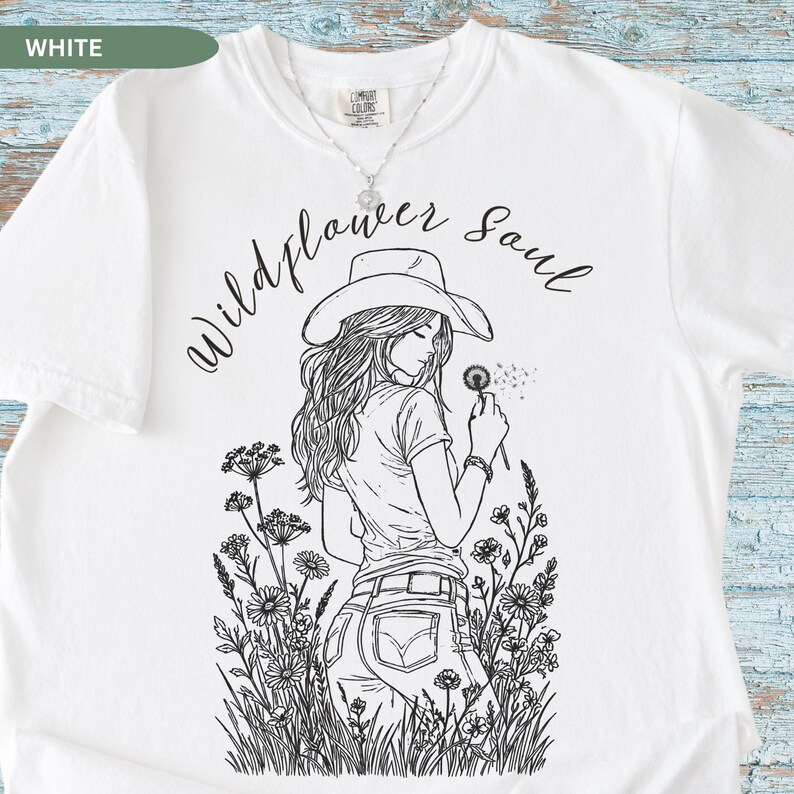 Puede incluir: Camiseta blanca con un gr&aacute;fico negro de una mujer con sombrero soplando un diente de le&oacute;n, rodeada de flores silvestres. El texto "Wildflower Soul" est&aacute; encima. La camiseta es de la marca Comfort Colors.