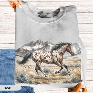May include: An ash gray crewneck sweatshirt featuring a graphic illustration of a spotted horse galloping through a field with tall grasses and shrubs, set against a backdrop of snow-capped mountain peaks under a clear sky.