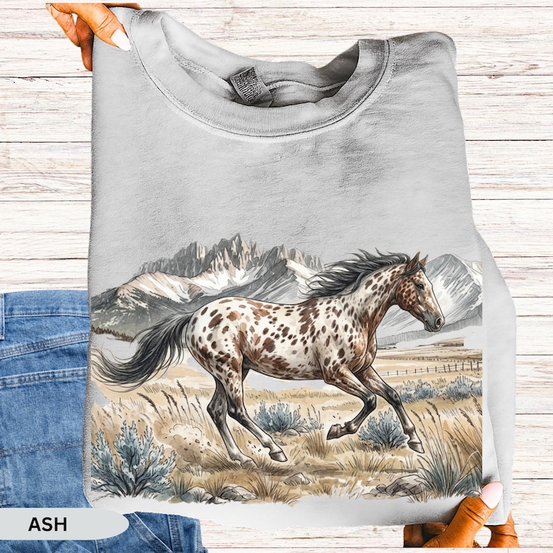 May include: An ash gray crewneck sweatshirt featuring a graphic illustration of a spotted horse galloping through a field with tall grasses and shrubs, set against a backdrop of snow-capped mountain peaks under a clear sky.