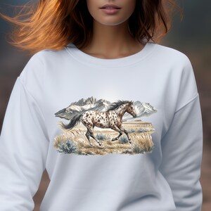 May include: A white long-sleeve sweatshirt featuring a detailed graphic print of a spotted Appaloosa horse galloping through a grassy field. The background depicts a mountain range under a bright sky, rendered in a sketch-like artistic style.