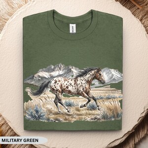May include: A military green crew neck t-shirt featuring a graphic illustration of a spotted horse galloping through a field with mountains in the background. The design includes muted tones of brown, white, and blue, with a Gildan brand label at the neck.
