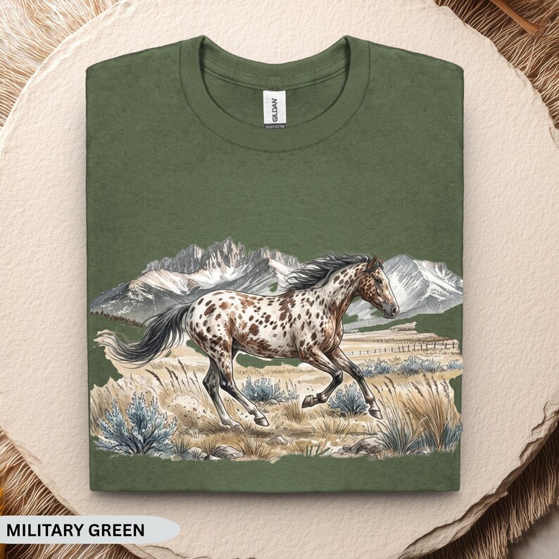 May include: A military green crew neck t-shirt featuring a graphic illustration of a spotted horse galloping through a field with mountains in the background. The design includes muted tones of brown, white, and blue, with a Gildan brand label at the neck.
