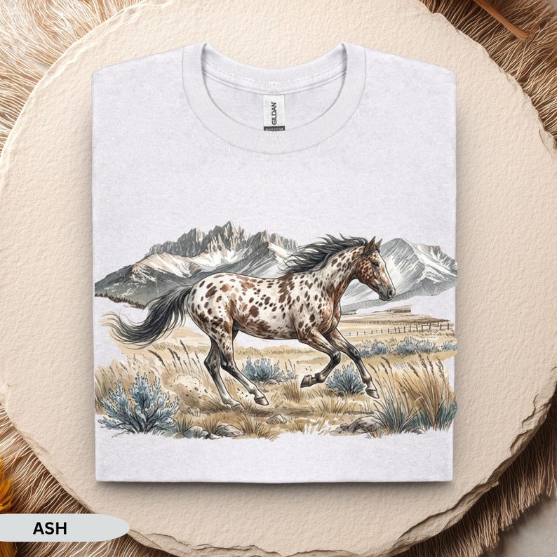 May include: An ash gray crew neck t-shirt featuring a graphic illustration of a brown and white spotted horse galloping through a grassy field with mountains in the background. The Gildan brand tag is visible at the inner neckline of the shirt.