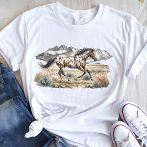 May include: A white short-sleeve t-shirt featuring a graphic print of a brown and white spotted horse galloping through a grassy field with mountains in the background. The horse has a dark mane and tail, and the landscape includes muted earth tones.