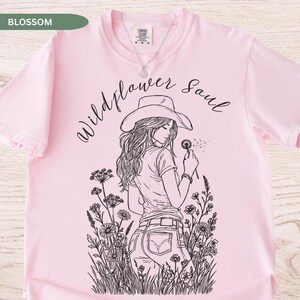 Puede incluir: Camiseta rosa claro con la frase "Wildflower Soul" y una ilustraci&oacute;n en blanco y negro de una mujer con sombrero de vaquero sosteniendo un diente de le&oacute;n. La camiseta es de la marca Comfort Colors.
