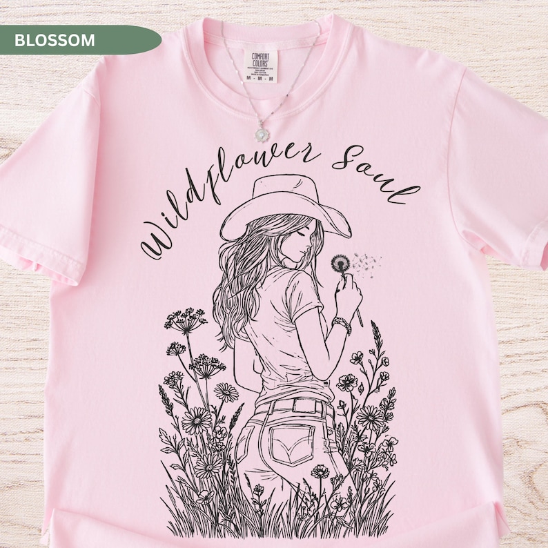 Puede incluir: Camiseta rosa claro con la frase "Wildflower Soul" y una ilustraci&oacute;n en blanco y negro de una mujer con sombrero de vaquero sosteniendo un diente de le&oacute;n. La camiseta es de la marca Comfort Colors.