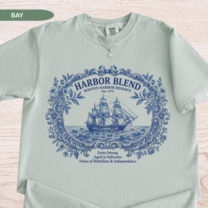 May include: A light sage green short-sleeve t-shirt featuring a dark blue graphic print of a sailing ship within an ornate floral frame. The text reads Harbor Blend Boston Harbor Reserve Est. 1773 Extra Strong Aged in Saltwater Notes of Rebellion & Independence.