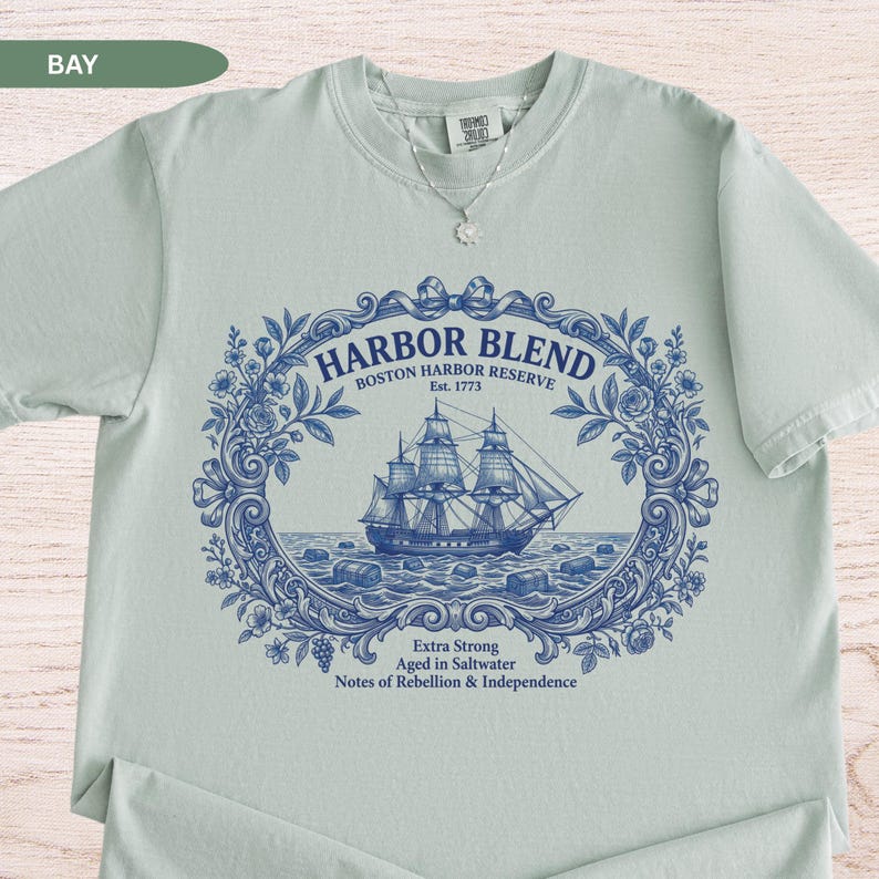 May include: A light sage green short-sleeve t-shirt featuring a dark blue graphic print of a sailing ship within an ornate floral frame. The text reads Harbor Blend Boston Harbor Reserve Est. 1773 Extra Strong Aged in Saltwater Notes of Rebellion & Independence.