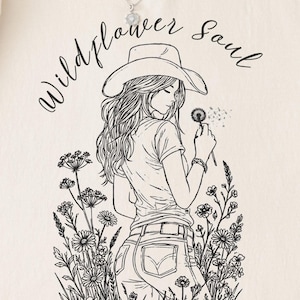 Puede incluir: Sudadera color crema con una ilustraci&oacute;n en blanco y negro de una mujer con sombrero de vaquero sosteniendo un diente de le&oacute;n. El texto "Wildflower Soul" est&aacute; arqueado sobre la ilustraci&oacute;n.