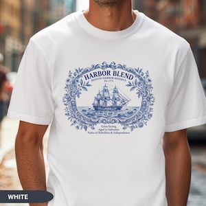 May include: A white short-sleeve t-shirt featuring a blue graphic print of a sailing ship inside an ornate floral frame. The text reads Harbor Blend, Boston Harbor Reserve, Est. 1773, Extra Strong, Aged in Saltwater, Notes of Rebellion & Independence.