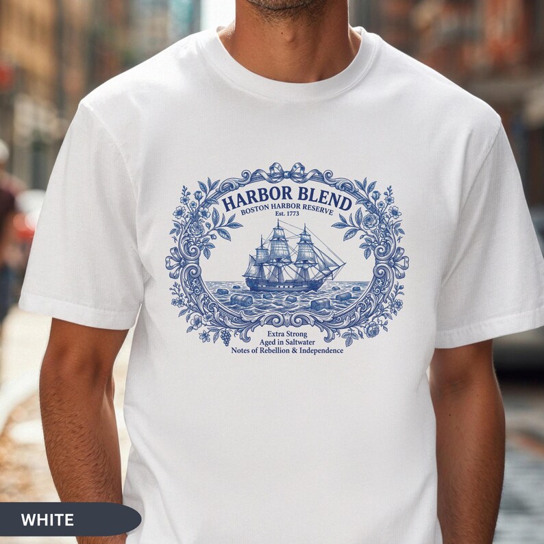 May include: A white short-sleeve t-shirt featuring a blue graphic print of a sailing ship inside an ornate floral frame. The text reads Harbor Blend, Boston Harbor Reserve, Est. 1773, Extra Strong, Aged in Saltwater, Notes of Rebellion & Independence.