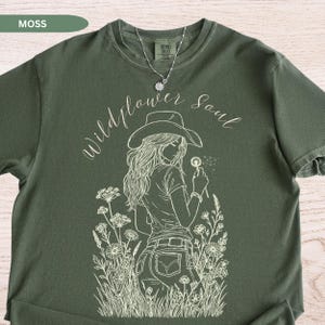 Puede incluir: Camiseta verde musgo con un gr&aacute;fico blanco de una mujer con sombrero de vaquero sosteniendo un diente de le&oacute;n. El texto "Wildflower Soul" est&aacute; encima del gr&aacute;fico. Se ve un collar plateado. La camiseta es de estilo casual.