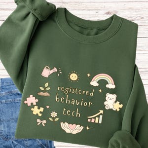 Op de afbeelding: Donkergroene sweatshirt met de tekst "registered behavior tech" omringd door speelse illustraties. Het ontwerp bevat een gieter, zon, regenboog, beer, puzzelstukjes en een groeiende plant.