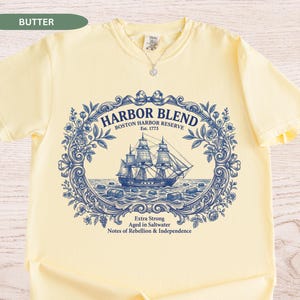 May include: A butter yellow t-shirt featuring a dark blue graphic print of a sailing ship inside an ornate floral frame. The text reads HARBOR BLEND BOSTON HARBOR RESERVE Est. 1773 Extra Strong Aged in Saltwater Notes of Rebellion & Independence.