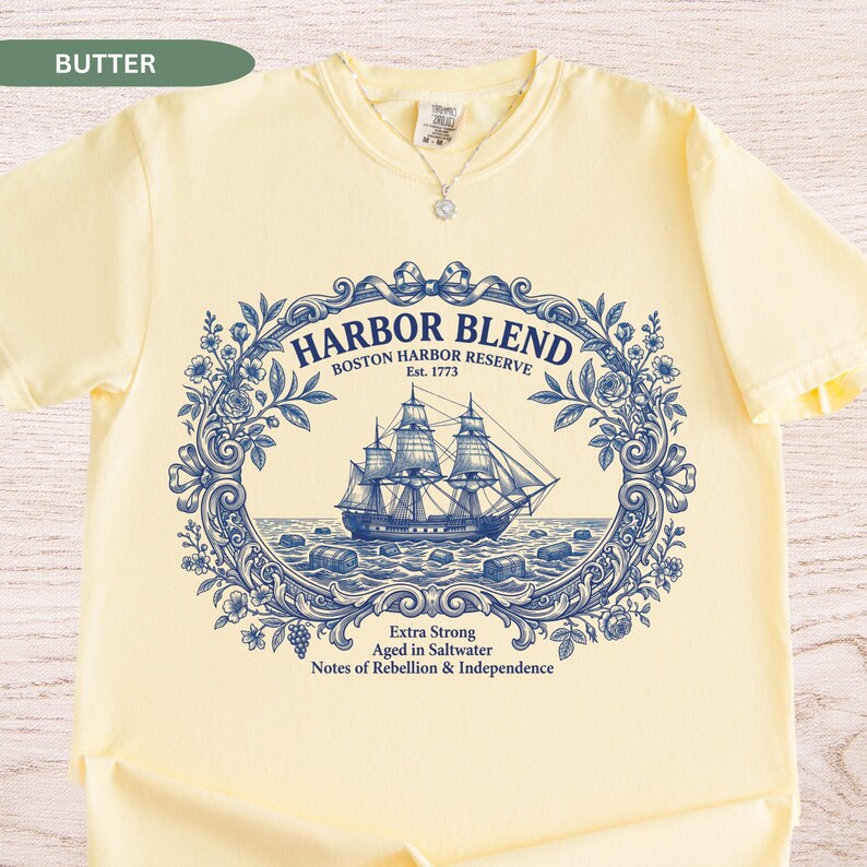 May include: A butter yellow t-shirt featuring a dark blue graphic print of a sailing ship inside an ornate floral frame. The text reads HARBOR BLEND BOSTON HARBOR RESERVE Est. 1773 Extra Strong Aged in Saltwater Notes of Rebellion & Independence.