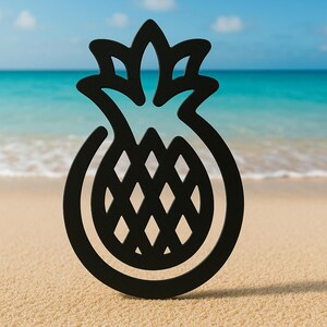 May include: A black pineapple-shaped decorative object stands on a sandy beach with the ocean in the background. The pineapple design features a detailed diamond pattern and a leafy crown, evoking a tropical theme.
