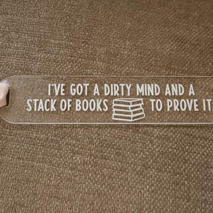 Puede incluir: Marcapáginas transparente de acrílico con esquinas redondeadas y un agujero para una cinta. El texto blanco dice "I'VE GOT A DIRTY MIND AND A STACK OF BOOKS TO PROVE IT", con un dibujo de una pila de libros. Una cinta rosa está adjunta.