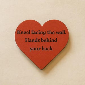 Może przedstawiać: Czerwony przedmiot w kształcie serca z czarnym napisem "Kneel facing the wall. Hands behind your back". Obiekt znajduje się na neutralnym tle.