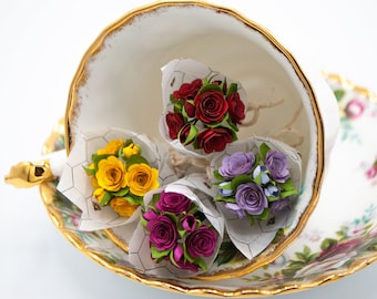 May include: Four small paper flower bouquets in a teacup. The bouquets are red, yellow, purple, and pink. The teacup is white with gold trim and is sitting on a saucer with a floral pattern.
