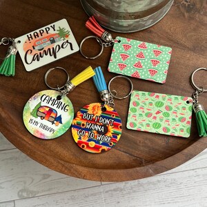 May include: A collection of keychains with various designs and colorful tassels. The keychains feature camping themes, watermelon slices, and phrases like "Happy Camper" and "But I don't wanna go to work". They are arranged on a wooden tray.