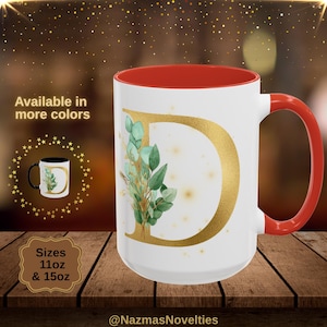 May include: White ceramic mug with a red interior and handle, featuring a gold letter "D" and a botanical design. The mug is available in 11oz and 15oz sizes. The text "Available in more colors" is displayed.