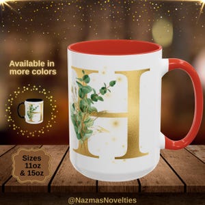 Puede incluir: Taza de cerámica blanca con la letra "H" dorada y detalles botánicos verdes. El interior y el asa son rojos. Disponible en tamaños de 11 oz y 15 oz. La taza está decorada con un diseño floral dorado.