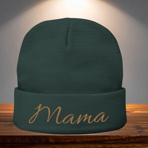 May include: A dark green beanie with the word "Mama" embroidered in a gold script font across the folded brim. The hat is made of a soft, knitted material.