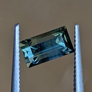 Peut inclure: Une pierre précieuse rectangulaire de couleur teal, tenue par une pince argentée. La gemme a une taille à facettes, affichant un mélange de teintes bleues et vertes. Le fond est marron neutre.
