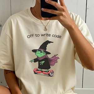 May include: Cream-colored t-shirt with the text "Off to write code." The graphic features a green frog wearing a witch hat, scarf, and robe, riding a red hoverboard. The frog is facing left.