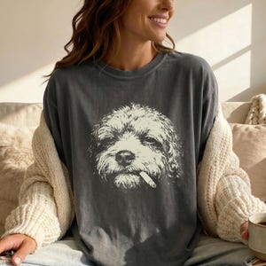 May include: Dark grey t-shirt featuring a white illustration of a dog with a cigarette in its mouth. The dog's face is detailed with fur texture. The shirt is a relaxed fit, suitable for casual wear.
