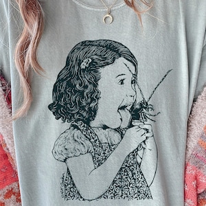 May include: A light gray t-shirt featuring a black and white illustration of a girl with her mouth open, seemingly eating a large insect. The graphic is detailed and artistic, with the artist's signature at the bottom.
