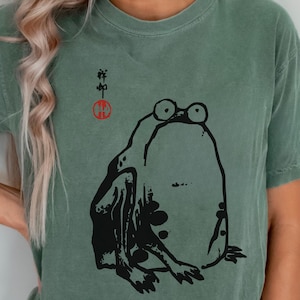 Könnte beinhalten: Salbeigrünes T-Shirt mit einer schwarzen Frosch-Illustration mit großen Augen. Das T-Shirt hat auch japanischen Text und ein rotes Kreissymbol. Ein lässiges, bequemes T-Shirt.