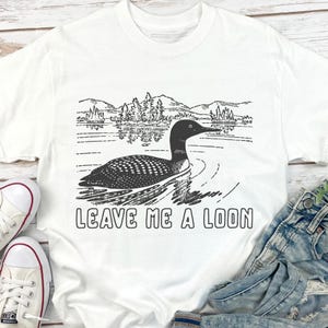 May include: White t-shirt with a black and white graphic of a loon swimming on a lake, with trees and mountains in the background. The text "LEAVE ME A LOON" is printed below the image.
