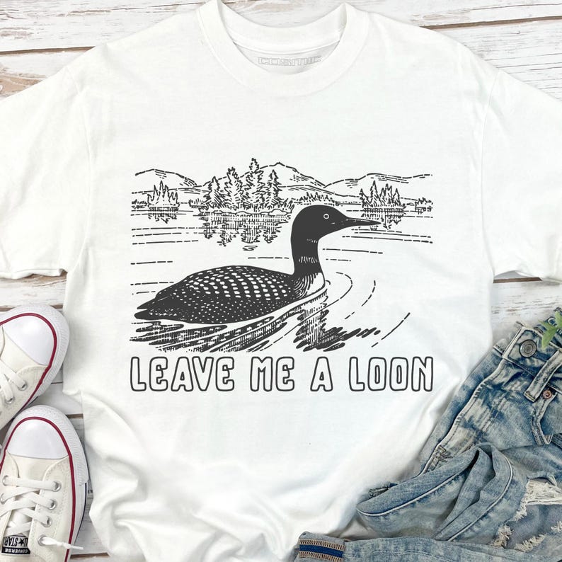 May include: White t-shirt with a black and white graphic of a loon swimming on a lake, with trees and mountains in the background. The text "LEAVE ME A LOON" is printed below the image.