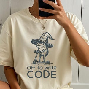 Op de afbeelding: Crèmekleurig T-shirt met een illustratie van een kikker met een tovenaars hoed op een scooter. De tekst "Off to write CODE" staat eronder in donkergrijs. Het T-shirt is een casual unisex stijl.