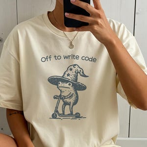 May include: Cream-colored t-shirt with the text "Off to write code." The graphic features a frog wearing a wizard hat and riding a scooter. The frog is gray, and the hat has star details.